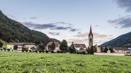 Vintl im Pustertal Das Bild zeigt eine grüne Wiese im Vordergrund mit einer Kirche und mehreren Häusern im Hintergrund. Die Kirche hat einen markanten roten Kirchturm, und das Dorf ist von bewaldeten Hügeln umgeben, während der Himmel leicht bewölkt ist.