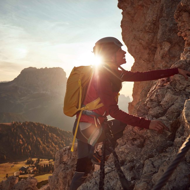 Summer holidays in South Tyrol: hiking and more The picture shows a person climbing a steep rock wall. The sun shines directly behind them, creating a dramatic silhouette. The person is wearing a helmet and a yellow backpack, while a wide mountain landscape can be seen in the background.