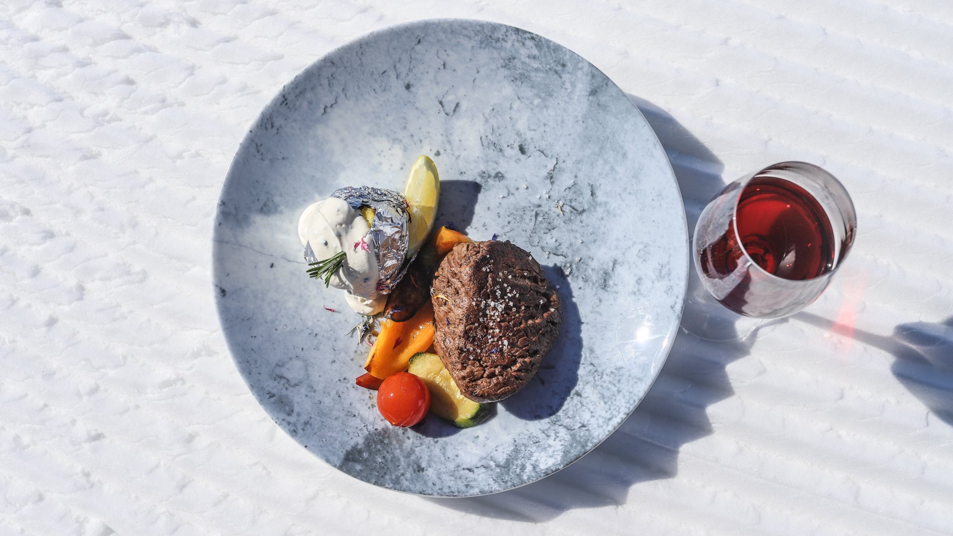 Rund um Meransen: Hütten und Almen Auf dem Bild ist ein appetitlich angerichteter Teller mit einem gegrillten Steak, begleitet von gegrilltem Gemüse und einer in Alufolie gewickelten Ofenkartoffel mit Kräutercreme, serviert auf einem marmorierten Teller auf einem schneebedeckten Untergrund neben einem Glas Rotwein.