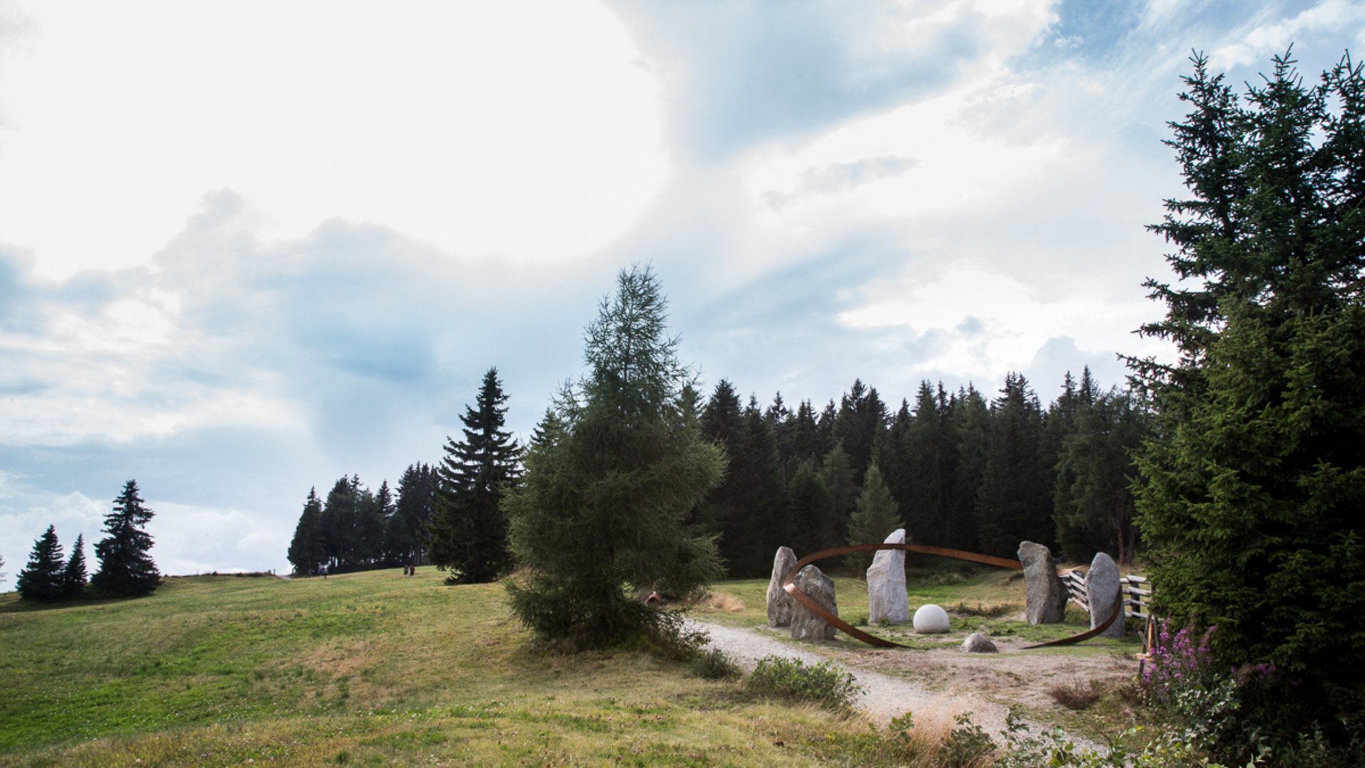 Der Schöpfungsweg in Rodeneck Das Bild zeigt eine grasbewachsene Landschaft mit vereinzelten Bäumen im Vordergrund und einem dichten Wald im Hintergrund. Auf einer Lichtung befindet sich eine Installation aus mehreren großen Steinen, die von einem metallischen Ring umgeben sind.