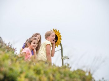 The mountain village of Meransen The picture shows three laughing children leaning against a large, yellow sunflower. The sunflower appears to be part of an installation or playground, and the children seem to be having fun while interacting with it.