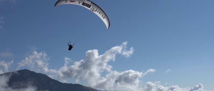 The Gitschberg webcams The picture shows a paraglider soaring high above a mountain landscape. The sky is clear and blue, with a few scattered clouds, and you can see the silhouette of a mountain in the background.