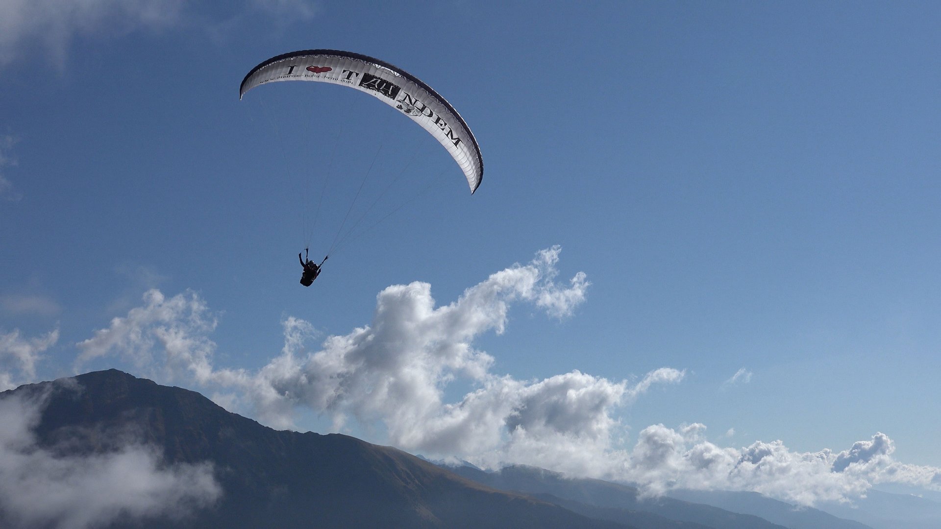 Paragliding v Jižním Tyrolsku Obrázek ukazuje paraglidingového letce, který vysoko nad horskou krajinou plachtí. Obloha je čistá a modrá, s několika roztroušenými mraky, a v pozadí je silueta hory.