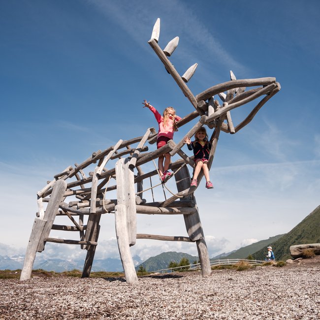 Euer Familienurlaub in Südtirol Zwei Kinder spielen auf einem großen, hölzernen Rentier-Spielplatz bei sonnigem Wetter
