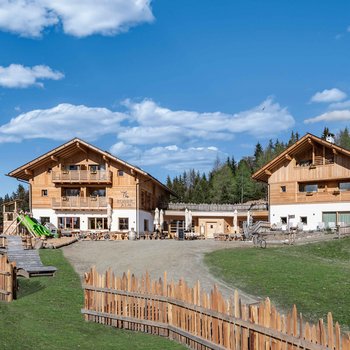 Südtiroler Almkost Das Bild zeigt zwei moderne Holzgebäude der "Roner Alm" unter einem klaren blauen Himmel mit vereinzelten weißen Wolken. Die Gebäude sind von einem Holzzaun umgeben und im Vordergrund befindet sich ein Spielplatz mit einer Rutsche sowie einige Holzbänke. Im Hintergrund sind Wälder und Hügel zu sehen.