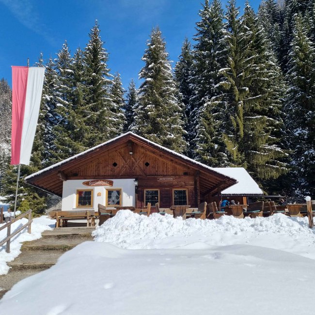 Kurzkofelhütte De foto toont een berghut met een houten gevel, waarvoor een Oostenrijkse vlag wappert. De hut is omgeven door diepe sneeuw en op de achtergrond staan met sneeuw bedekte naaldbomen onder een heldere, blauwe lucht.