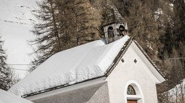 Pfunders: urig und idyllisch Das Bild zeigt eine kleine, schneeweiße Kapelle mit einem schneebedeckten Dach. Der Glockenturm aus Holz sitzt auf dem Dach, und im Hintergrund sind schneebedeckte Bäume und Hänge zu sehen.