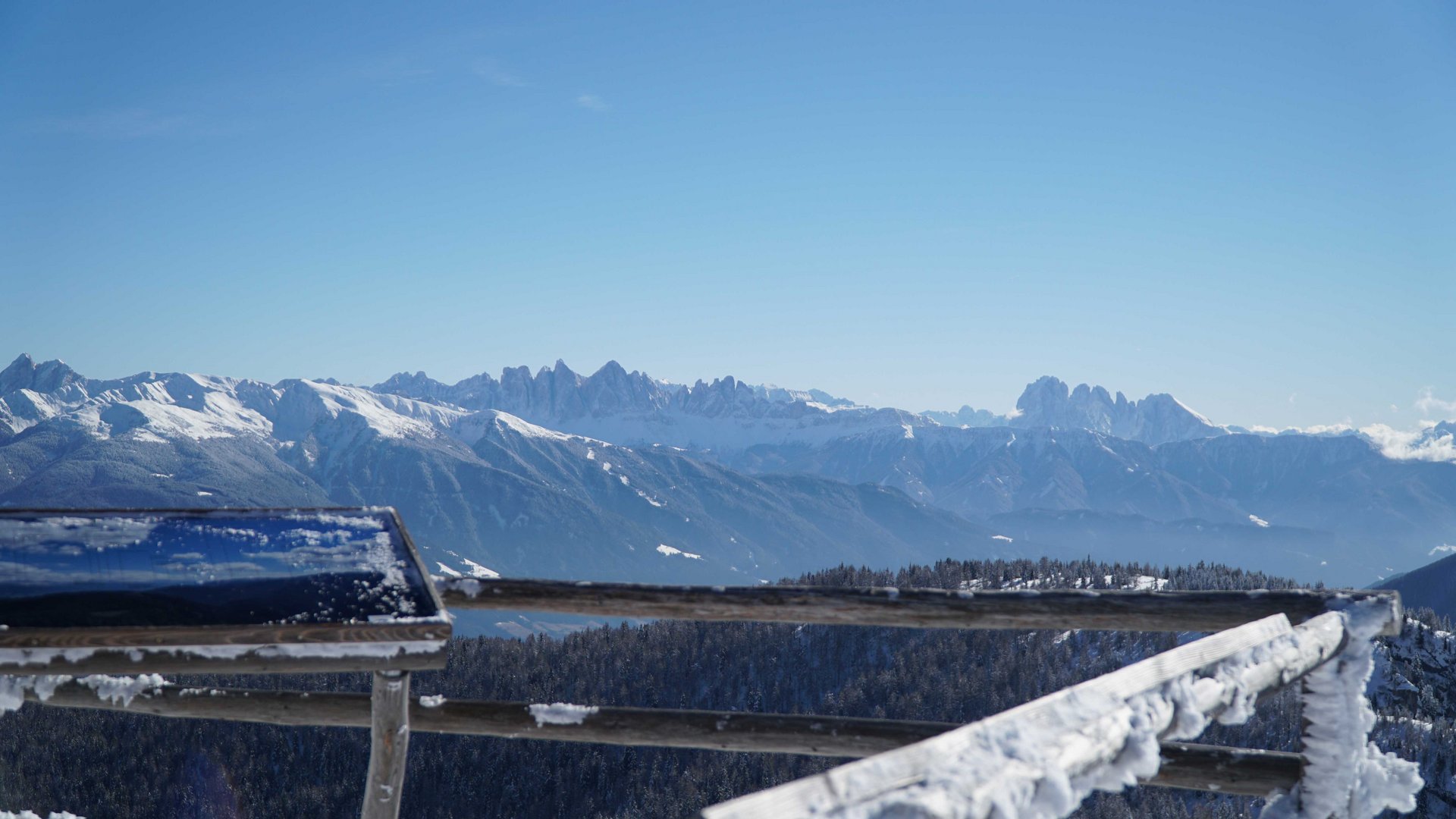 Vals - Jochtal - Steinermandl The image shows a winter mountain landscape with snow-covered peaks in the background under a bright blue sky. In the foreground, there is a snow-covered railing and a slightly blurred sign or information board.