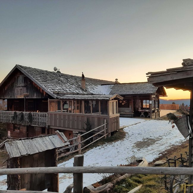 Pertinger Alm The picture shows a rustic wooden mountain hut in winter at sunset. The hut has a large balcony and is partially covered with snow. In the background, there are coniferous trees and mountains, while the sun sets on the horizon.