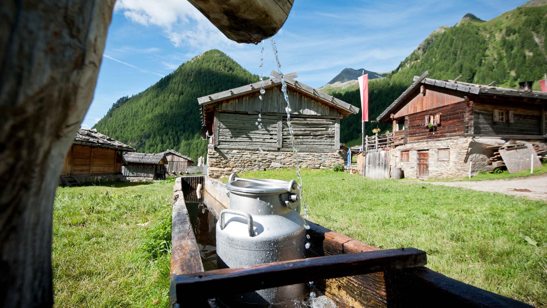 Fane Alm – South Tyrol’s most idyllic Alpine village The picture shows a traditional wooden well from which water flows into a trough. In the foreground stands a silver milk can in the water, while in the background several rustic wooden huts can be seen in front of a green mountain landscape.