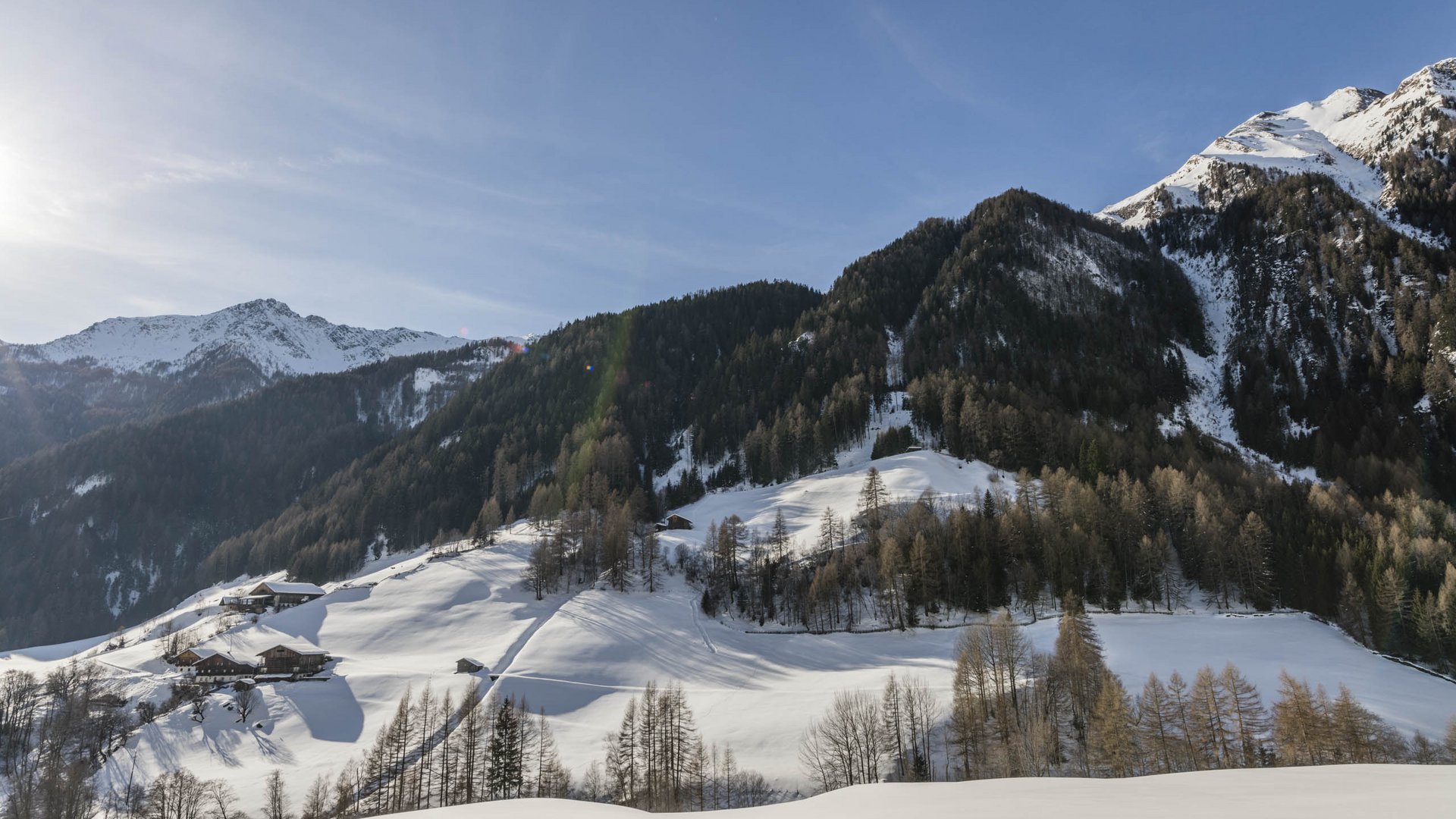 Pfunders: urig und idyllisch Das Bild zeigt eine schneebedeckte Berglandschaft mit mehreren Häusern, die an den Hängen verteilt sind. Die Berge im Hintergrund sind teilweise mit Wald bedeckt, und die Sonne scheint am klaren blauen Himmel, was lange Schatten auf den Schnee wirft.