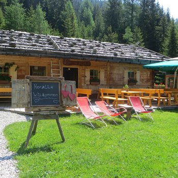Südtiroler Almkost Das Bild zeigt eine charmante, rustikale Berghütte aus Holz mit einem begrüßenden Schild "Herzlich Willkommen" davor, umgeben von grünen Bäumen und einem entspannten Sitzbereich mit Holztischen, Bänken und Liegestühlen unter einem Sonnenschirm.