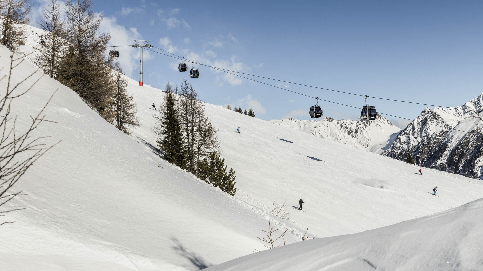 Lyžařská oblast Gitschberg Jochtal Obrázek zobrazuje zasněžený lyžařský svah s několika lyžaři sjíždějícími kopec dolů. Nad ním vede lanovka s gondolami, která přepravuje lyžaře nahoru na horu. V pozadí jsou vidět zasněžené hory a jasná modrá obloha.