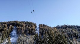The Altfasstal valley, Meransen The picture shows a cable car with gondolas passing over snow-covered trees and a wooded hill. The sky is clear and blue, and the cable car leads to a summit that is also covered with snow.