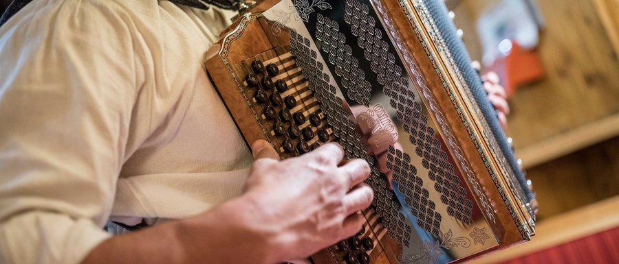 The Gitschberg webcams The picture shows a person playing a traditional accordion. The focus is on the hands operating the buttons and keys of the instrument, while the accordion features detailed decorations.