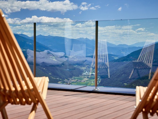 Willkommen in der Ferienregion Gitschberg Jochtal! Das Bild zeigt zwei Holz-Liegestühle auf einer Terrasse mit Glasgeländer, durch das man auf eine weitläufige Berglandschaft blickt. Die Aussicht zeigt grüne Täler, bewaldete Hügel und blaue Berge im Hintergrund unter einem leicht bewölkten Himmel.