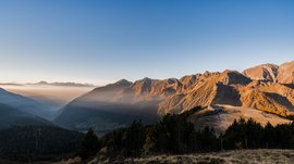 Pfunders: urig und idyllisch Das Bild zeigt eine weite Berglandschaft bei Sonnenaufgang oder Sonnenuntergang. Die Berge sind in warmes, goldenes Licht getaucht, während Nebel die Täler füllt. Am rechten Rand des Bildes ist eine kleine Berghütte zu sehen, die auf einem Hügel liegt und ebenfalls im sanften Licht erstrahlt.