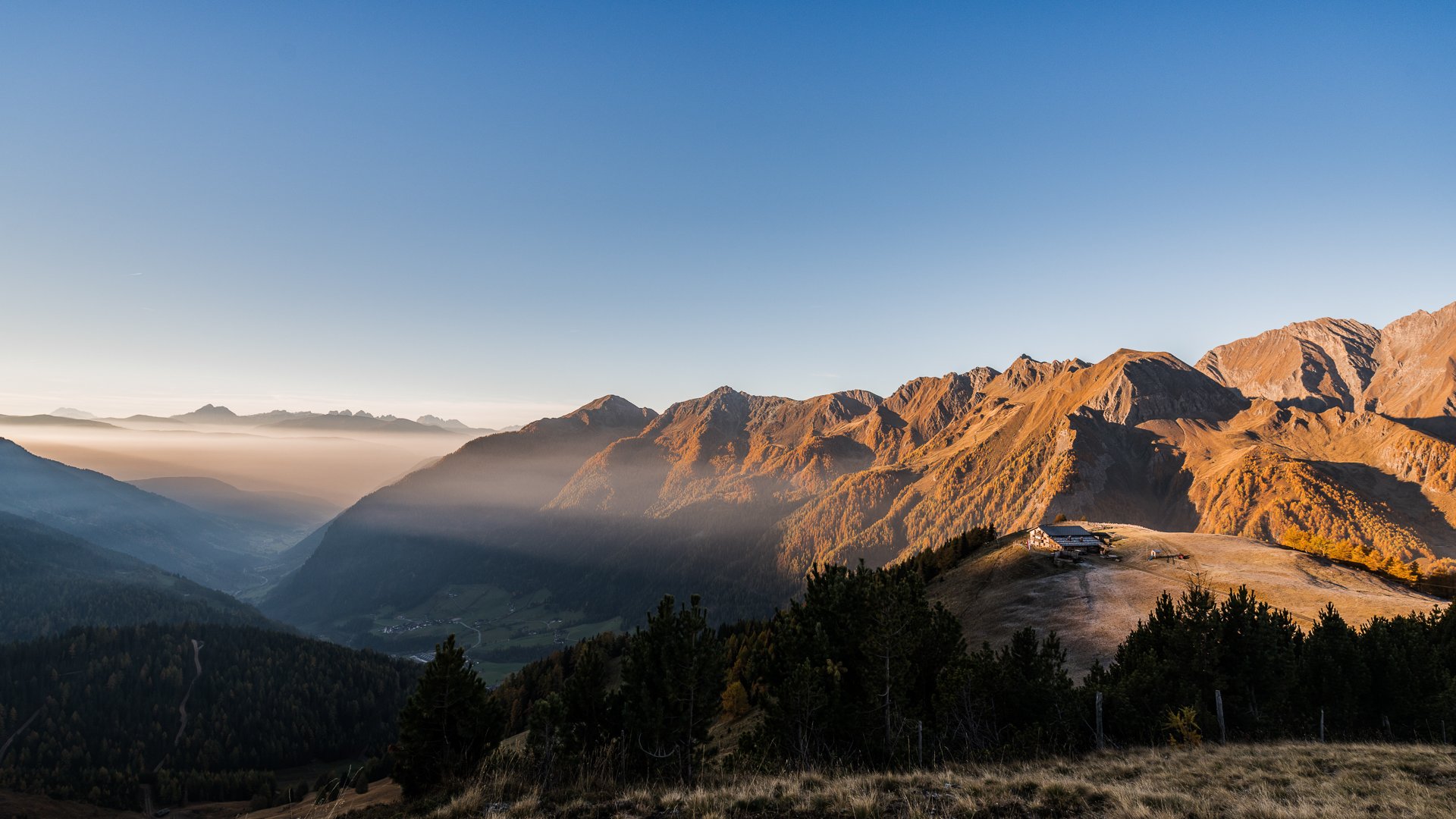 Pfunders: urig und idyllisch Das Bild zeigt eine weite Berglandschaft bei Sonnenaufgang oder Sonnenuntergang. Die Berge sind in warmes, goldenes Licht getaucht, während Nebel die Täler füllt. Am rechten Rand des Bildes ist eine kleine Berghütte zu sehen, die auf einem Hügel liegt und ebenfalls im sanften Licht erstrahlt.