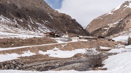 Fane Alm – South Tyrol’s most idyllic Alpine village The picture shows a winter mountain landscape with a small village surrounded by snow-covered and partly snow-free slopes. In the foreground a stream flows, and in the background some wooden huts and a small church can be seen.