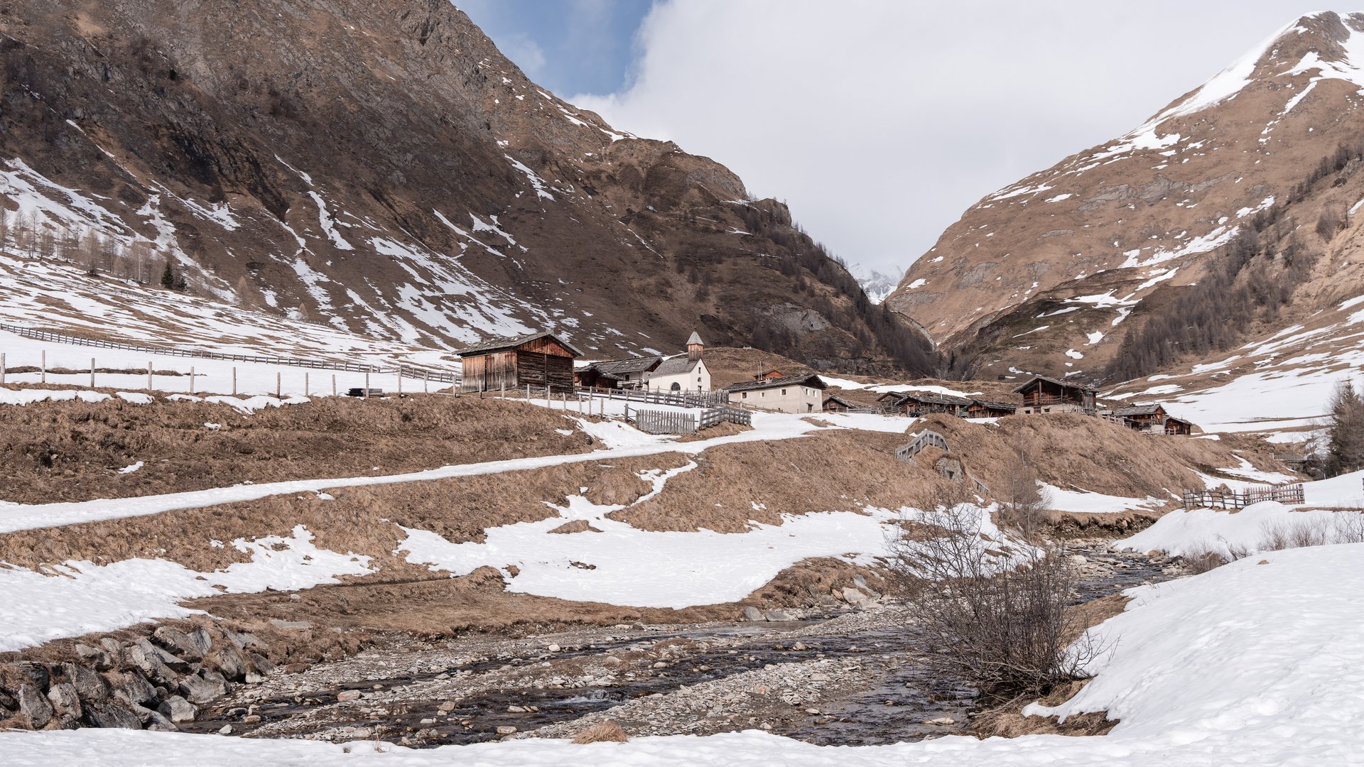 Fane Alm – South Tyrol’s most idyllic Alpine village The picture shows a winter mountain landscape with a small village surrounded by snow-covered and partly snow-free slopes. In the foreground a stream flows, and in the background some wooden huts and a small church can be seen.