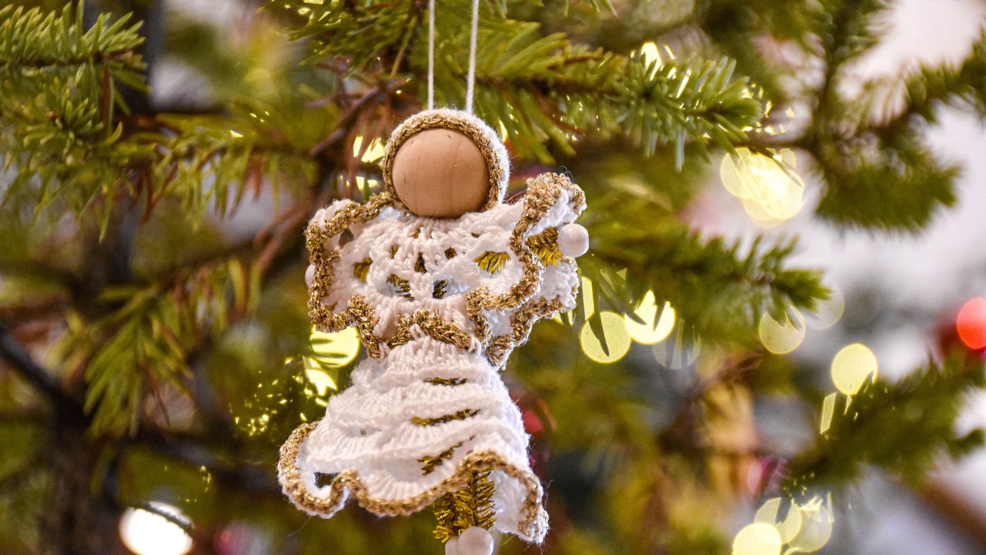 Advent in Terenten The image shows a handmade Christmas decoration in the shape of an angel, made of crocheted white fabric with golden embellishments, hanging on a Christmas tree. In the background, blurred lights and green fir branches glow.