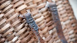 De Pfunderer Höfeweg De foto toont een close-up van een gevlochten mand gemaakt van houten strips. Twee roestige metalen banden die aan de mand zijn bevestigd, zijn op de voorgrond te zien, waarbij de structuur van de mand en de scheuren in het metaal duidelijk zichtbaar zijn.
