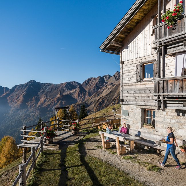 Gampiel Alm De foto toont een traditionele houten berghut, waar gasten buiten genieten van het zonnige uitzicht op herfstachtige bergen.