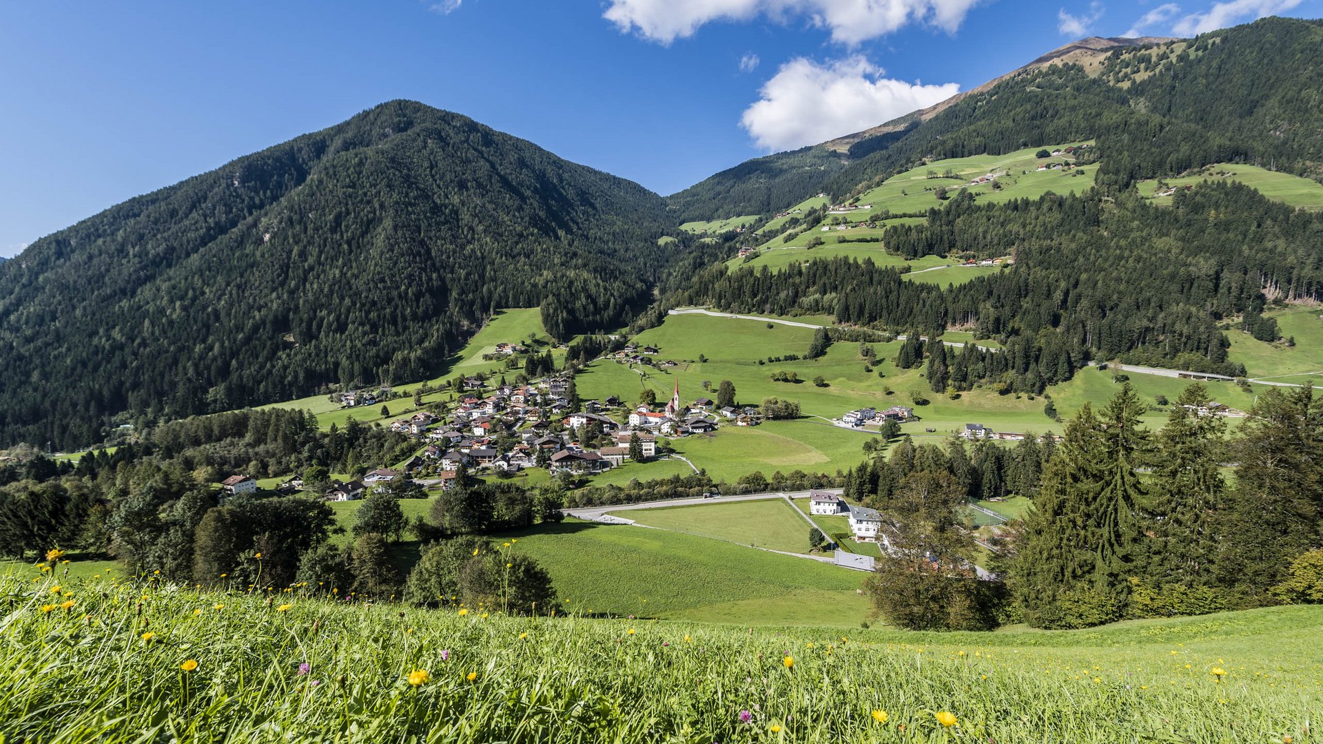Pfunders: urig und idyllisch Das Bild zeigt eine malerische Landschaft mit grünen Hügeln und einem kleinen Dorf, das in einem Tal eingebettet ist. Im Vordergrund sind blühende Wiesen, während der Hintergrund von bewaldeten Bergen und einem blauen Himmel mit ein paar Wolken dominiert wird.