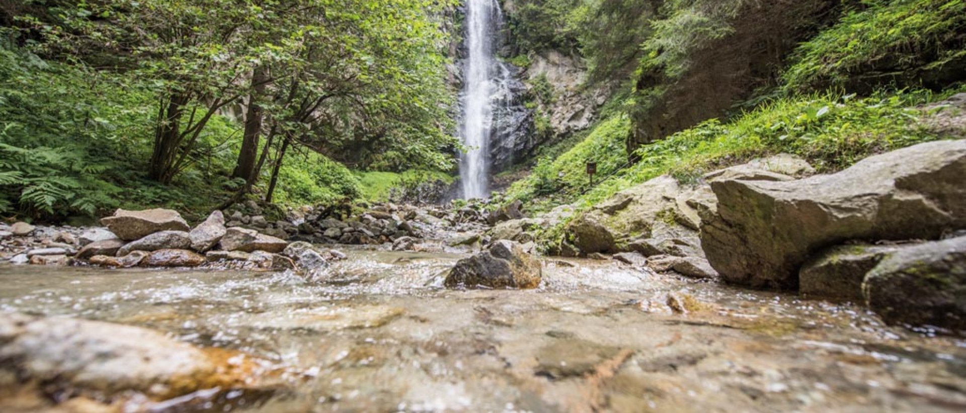 Výjimečně krásná: pěší turistika v Jižním Tyrolsku The picture shows a picturesque waterfall in the middle of a dense forest. In the foreground, a clear stream flows over stones, while the waterfall in the background plunges into the depths, surrounded by lush greenery.