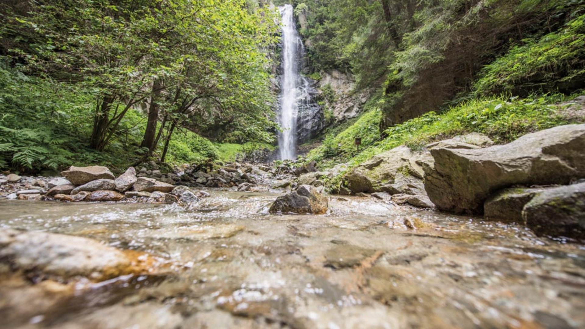 De Elfenweg bij Vintl De foto toont een schilderachtige waterval te midden van een dicht bos. Op de voorgrond stroomt een heldere beek over stenen, terwijl de waterval op de achtergrond in de diepte stort, omgeven door weelderig groen.