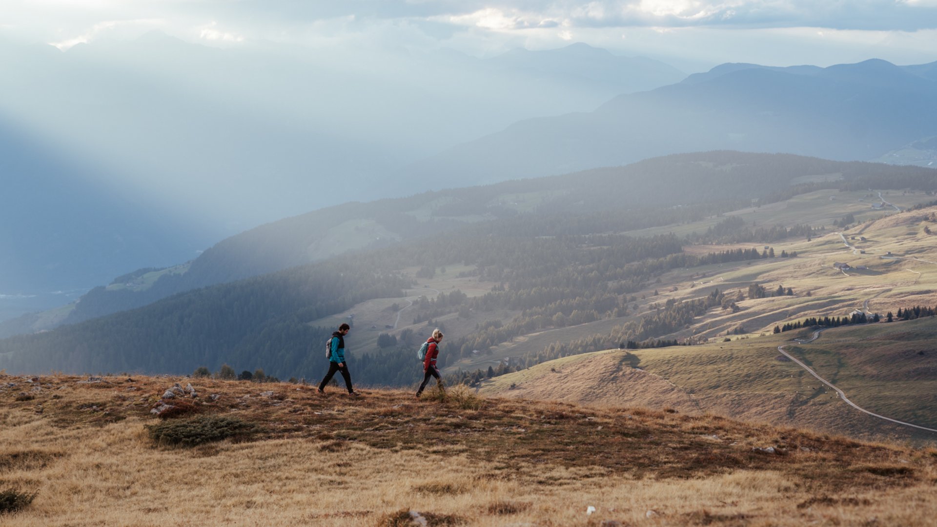 De mooiste wandelroutes in de Dolomieten De afbeelding toont twee wandelaars die over een met gras bedekte heuvel lopen. Op de achtergrond strekt zich een uitgestrekt, heuvelachtig landschap uit onder een bewolkte hemel, waar enkele zonnestralen dramatisch doorheen schijnen en de scène in zacht licht dompelen.