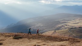 De mooiste wandelroutes in de Dolomieten De afbeelding toont twee wandelaars die over een met gras bedekte heuvel lopen. Op de achtergrond strekt zich een uitgestrekt, heuvelachtig landschap uit onder een bewolkte hemel, waar enkele zonnestralen dramatisch doorheen schijnen en de scène in zacht licht dompelen.