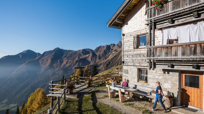 Gitschberg Jochtal: Ubytování a nabídky The picture shows a mountain hut on a sunny slope. People are sitting at a wooden table in front of the hut, and an impressive mountain landscape stretches out in the background under a bright blue sky.