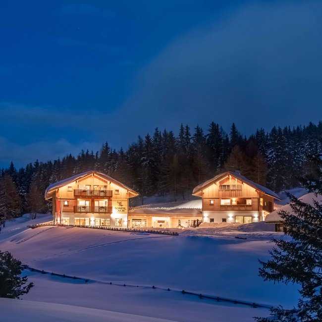 Naturhotel Runa De foto toont twee verlichte gebouwen van de "Roner Alm" in een besneeuwd landschap bij nacht. De gebouwen stralen warm licht uit en worden omgeven door met sneeuw bedekte bomen en heuvels, de lucht is donkerblauw en wolkenloos.