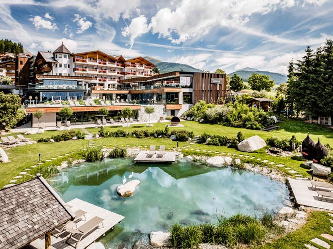 Willkommen in der Ferienregion Gitschberg Jochtal! Das Bild zeigt ein großes Hotelgebäude mit mehreren Etagen und Balkonen, umgeben von einer gepflegten Gartenanlage. Im Vordergrund befindet sich ein natürlicher Teich mit Liegen auf Holzplattformen, der von grünem Rasen und Steinen umgeben ist.