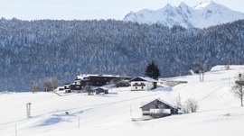 The mountain village of Meransen The picture shows a winter landscape with snow-covered hills and buildings, surrounded by a dense, snow-covered forest and tall, snow-covered mountains in the background. The houses stand scattered on the hills and are partly modern, partly traditional.