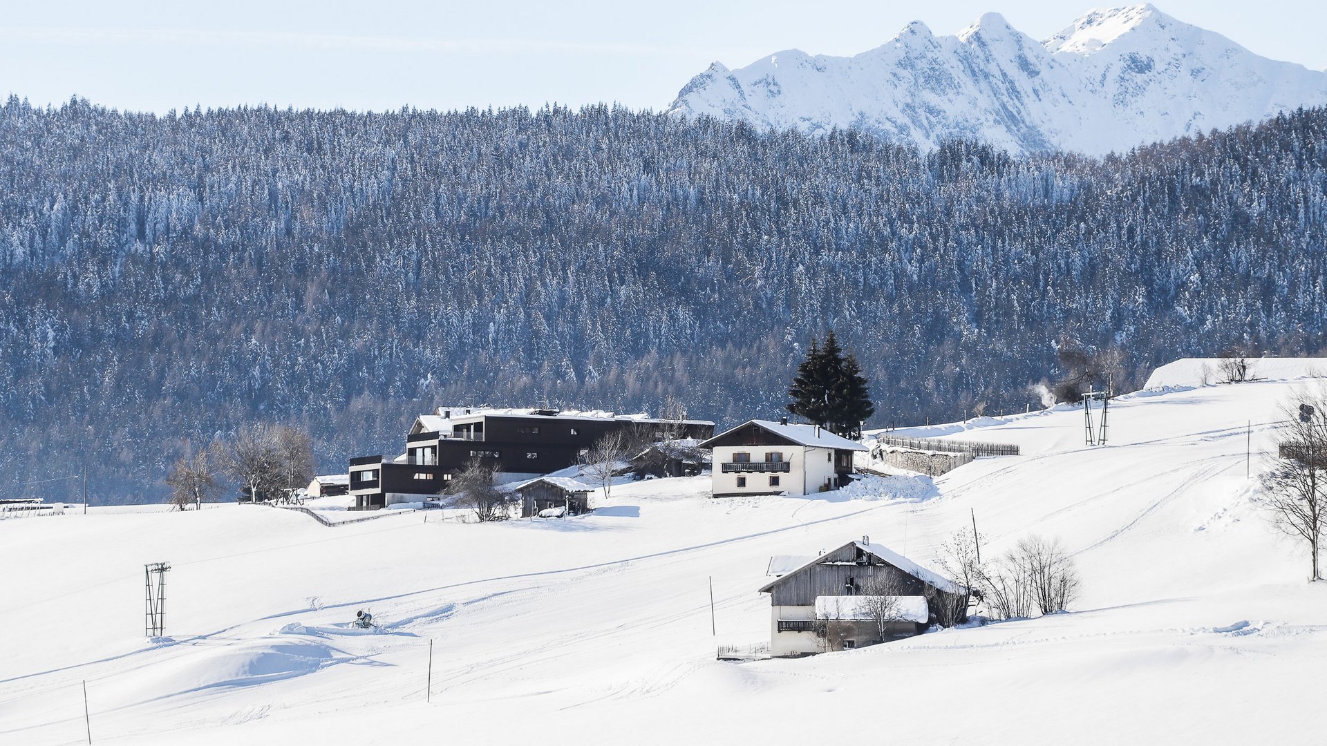 The mountain village of Meransen The picture shows a winter landscape with snow-covered hills and buildings, surrounded by a dense, snow-covered forest and tall, snow-covered mountains in the background. The houses stand scattered on the hills and are partly modern, partly traditional.