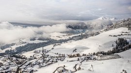 Skivakantie met kinderen in Zuid-Tirol/Terenten De foto toont een besneeuwd landschap met een klein dorp op de voorgrond en uitgestrekte heuvels op de achtergrond, bedekt met bossen en velden. Dichte wolken hangen boven de heuvels, terwijl op de achtergrond met sneeuw bedekte bergen te zien zijn.