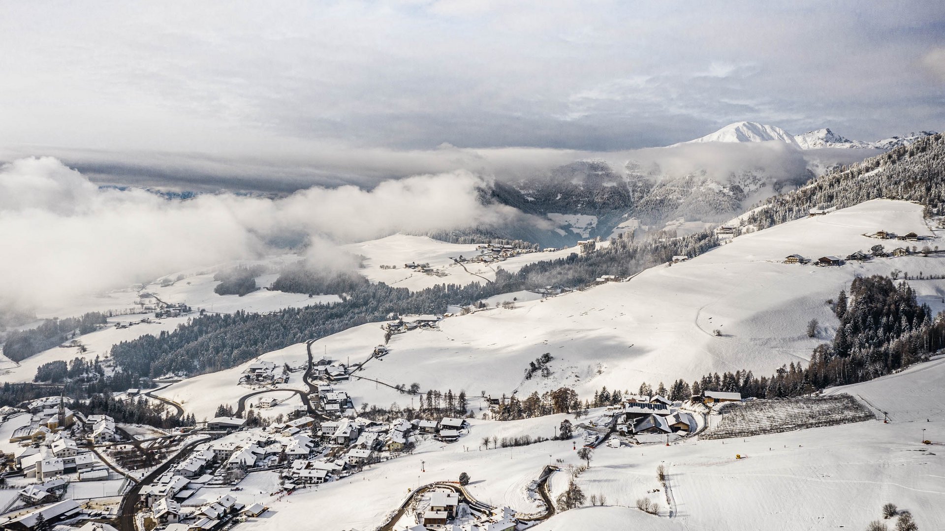 Skivakantie met kinderen in Zuid-Tirol/Terenten De foto toont een besneeuwd landschap met een klein dorp op de voorgrond en uitgestrekte heuvels op de achtergrond, bedekt met bossen en velden. Dichte wolken hangen boven de heuvels, terwijl op de achtergrond met sneeuw bedekte bergen te zien zijn.