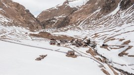 Fane Alm – South Tyrol’s most idyllic Alpine village The picture shows a small settlement of traditional wooden houses located in a valley, surrounded by snow-covered mountains. The snow is beginning to melt, revealing brown mountain surfaces, while a fence surrounds the area.