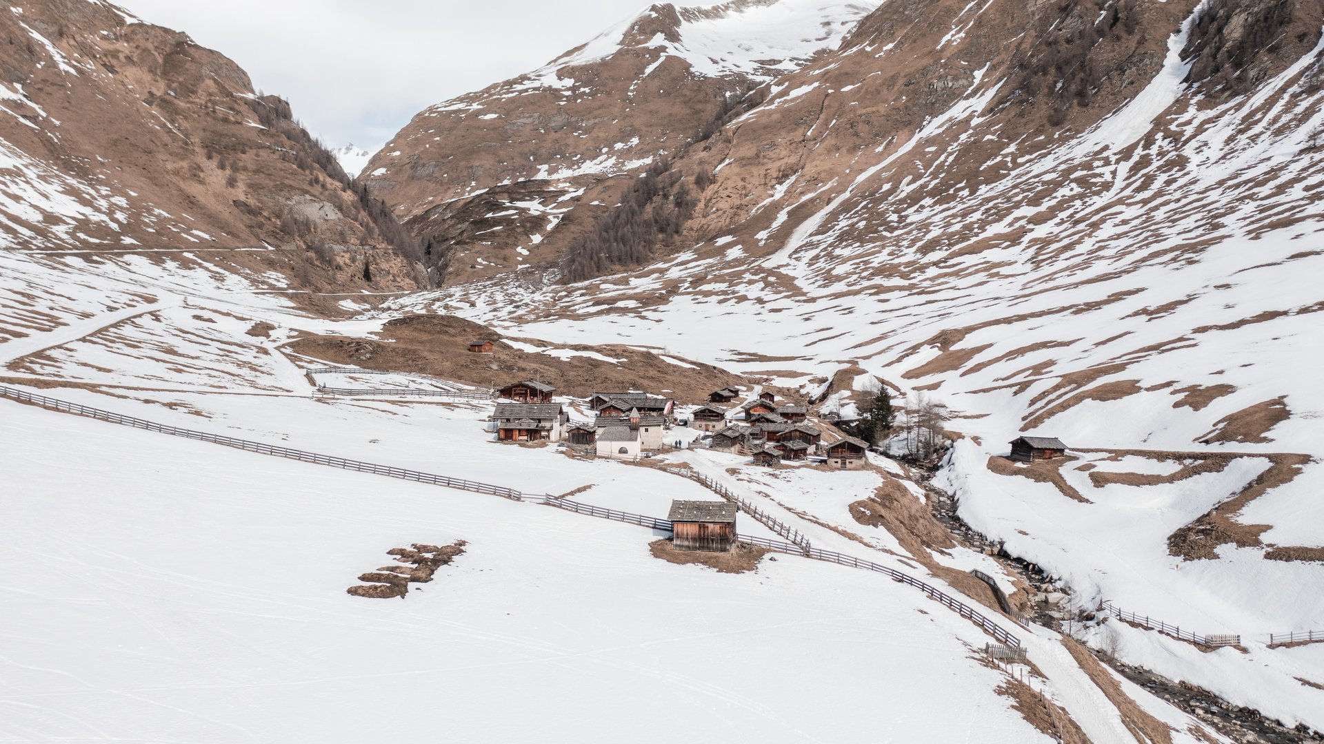 Fane Alm – South Tyrol’s most idyllic Alpine village The picture shows a small settlement of traditional wooden houses located in a valley, surrounded by snow-covered mountains. The snow is beginning to melt, revealing brown mountain surfaces, while a fence surrounds the area.