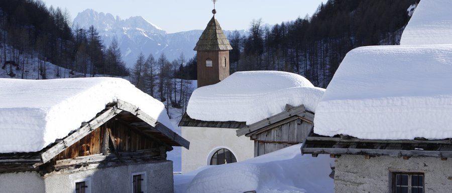 The Gitschberg webcams The picture shows a snowy mountain hut settlement with deep snow on the roofs of the buildings. In the background, there is a small church tower with a pointed roof, surrounded by snow-covered trees and mountains.