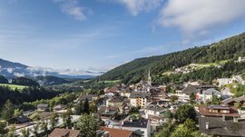 Mühlbach im Pustertal Das Bild zeigt eine kleine, malerische Stadt in einem grünen Tal, umgeben von bewaldeten Hügeln und Bergen. Im Zentrum des Bildes ragt ein Kirchturm mit einer Spitzhaube hervor, und die Gebäude sind von üppiger Vegetation und einigen verstreuten Wolken umgeben.