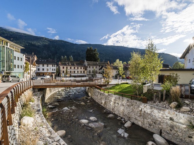 The Alpine hamlet of Spinges The picture shows a small bridge crossing a clear stream in a village. In the background, houses and green hills or mountains can be seen under a blue sky with few clouds.