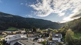 Vintl im Pustertal Das Bild zeigt eine kleine Stadt, eingebettet zwischen bewaldeten Hügeln, mit einigen Gebäuden im Vordergrund und einer Kirche mit einem hohen Turm in der Mitte. Die Sonne scheint über die Hügel und wirft lange Lichtstrahlen auf die Landschaft, während der Himmel mit einigen Wolken bedeckt ist.