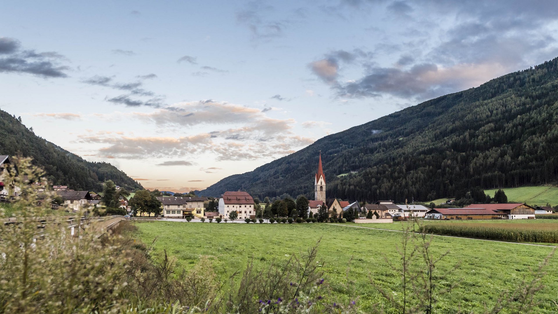 Vintl im Pustertal Das Bild zeigt eine weite grüne Wiese mit einem kleinen Dorf im Hintergrund, dessen Zentrum eine Kirche mit einem hohen spitzen Turm bildet. Umgeben ist das Dorf von bewaldeten Hügeln unter einem bewölkten Himmel bei Sonnenuntergang.