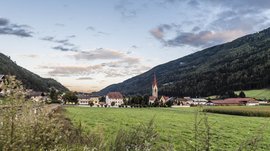 Vintl im Pustertal Das Bild zeigt eine weite grüne Wiese mit einem kleinen Dorf im Hintergrund, dessen Zentrum eine Kirche mit einem hohen spitzen Turm bildet. Umgeben ist das Dorf von bewaldeten Hügeln unter einem bewölkten Himmel bei Sonnenuntergang.