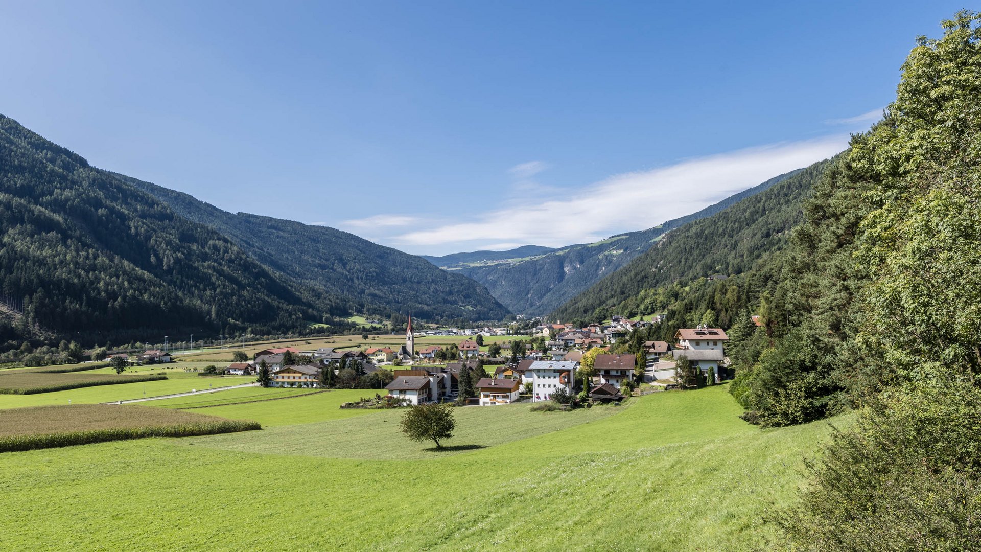 Vintl im Pustertal Das Bild zeigt ein grünes Tal mit einem kleinen Dorf im Vordergrund. In der Mitte des Dorfs steht eine Kirche mit einem hohen roten Turm, umgeben von Wohnhäusern und saftigen Wiesen, während bewaldete Hügel das Tal umschließen.