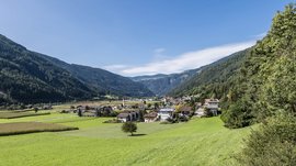 Vintl im Pustertal Das Bild zeigt ein grünes Tal mit einem kleinen Dorf im Vordergrund. In der Mitte des Dorfs steht eine Kirche mit einem hohen roten Turm, umgeben von Wohnhäusern und saftigen Wiesen, während bewaldete Hügel das Tal umschließen.