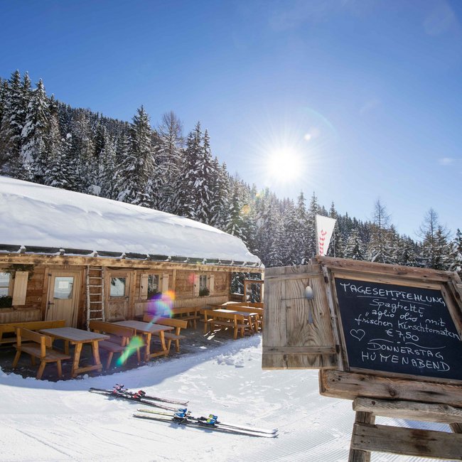 Bacherhütte Na obrázku je vidět zasněžená horská chata s dřevěnými lavicemi a stoly před chatou; vedle ní stojí tabule, která zobrazuje denní doporučení, zatímco v pozadí jsou zasněžené jehličnaté stromy a zářivě modrá obloha.